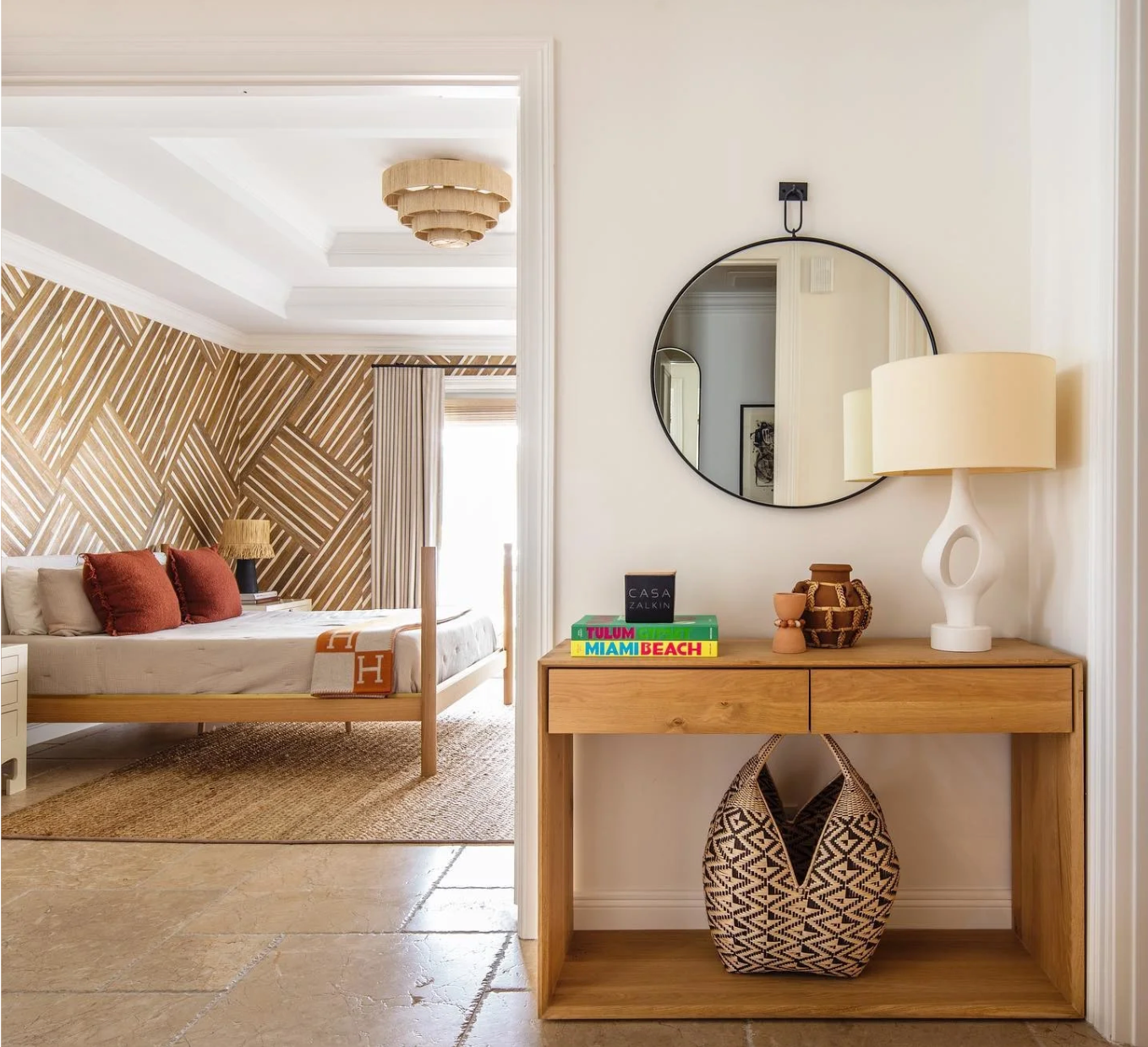 Modern bedroom and entryway with wood console, round mirror, white table lamp, and geometric accent wall.