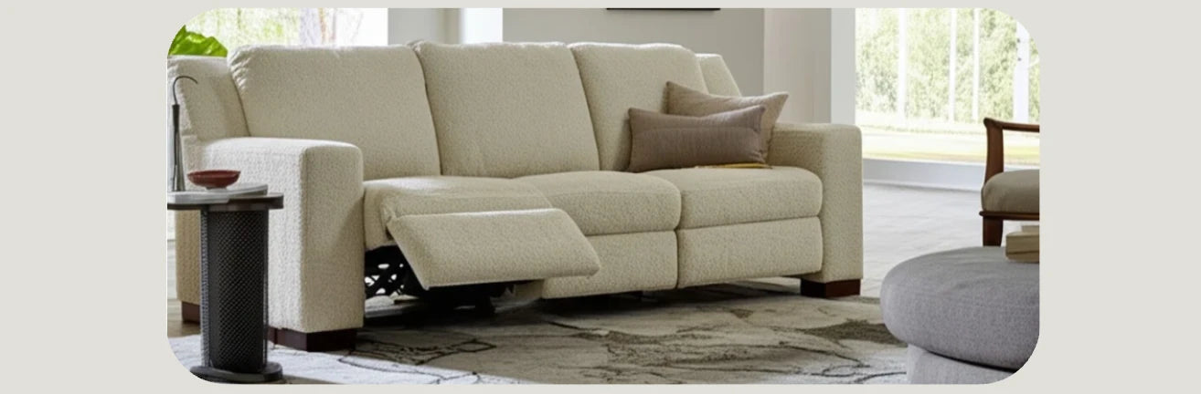Modern living room featuring a light-colored, three-seat reclining sofa with one extended footrest. The space includes a round side table, decorative pillows, and a neutral area rug, with natural light streaming in through large windows.