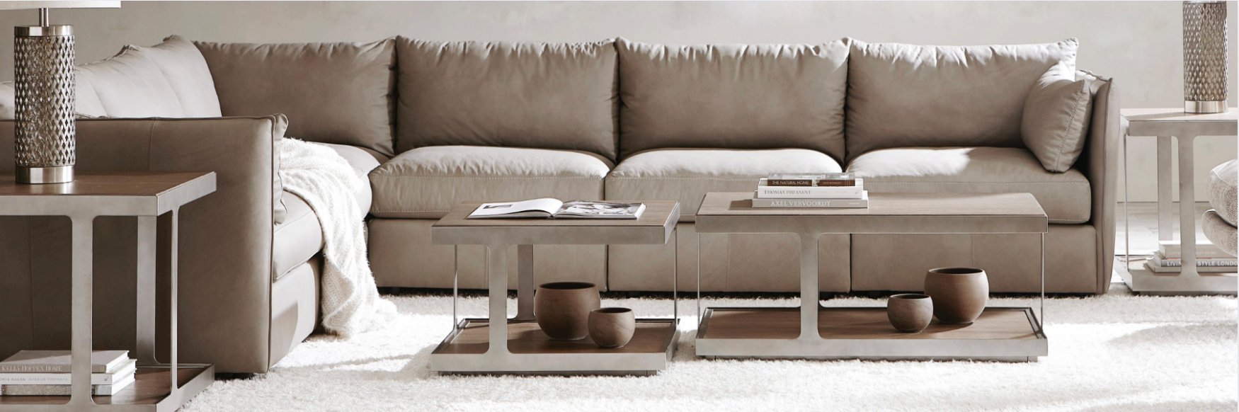 Modern living room with a large taupe sectional sofa, two matching side tables, and twin rectangular coffee tables styled with books and ceramic decor on a neutral rug