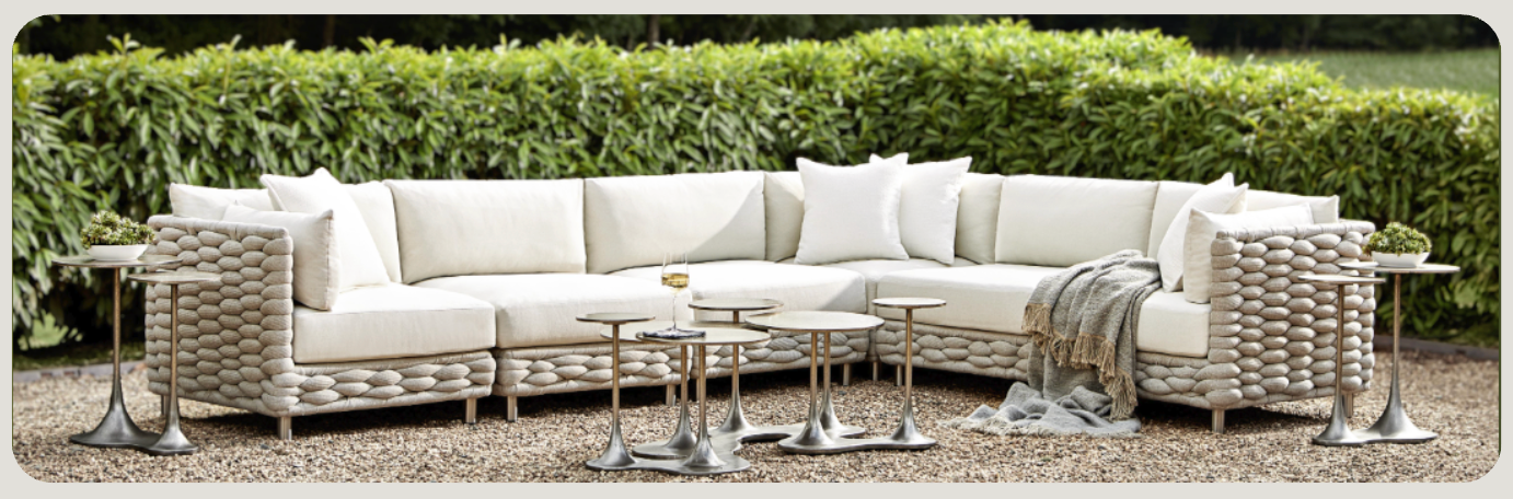 Outdoor patio setup featuring a large, L-shaped wicker sectional sofa with white cushions, surrounded by modern metal side tables and a soft throw blanket, set on a gravel surface with a lush green hedge in the background.