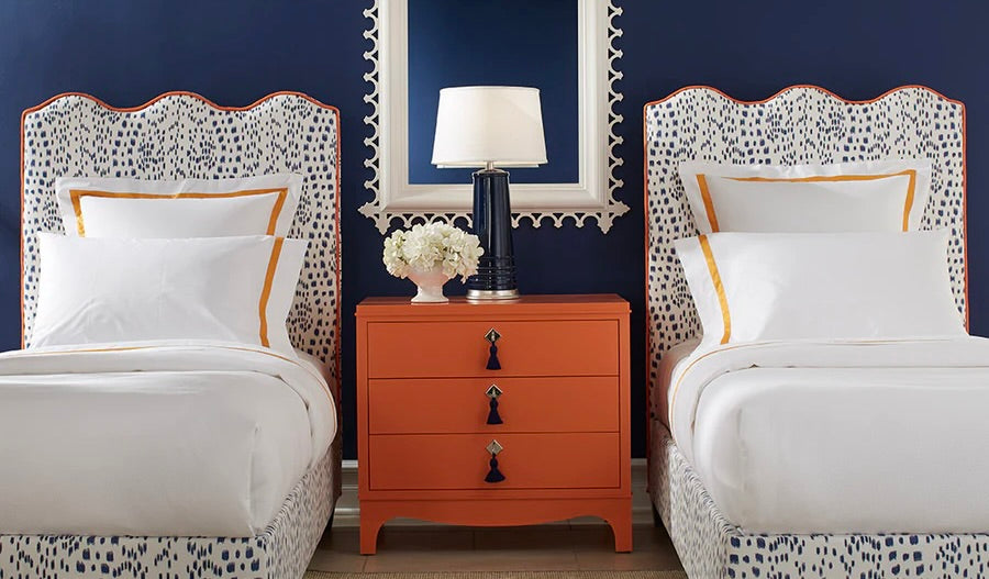 Twin beds with patterned upholstered headboards and white bedding, flanking an orange 3-drawer nightstand with navy lamp and flowers, against a navy blue wall