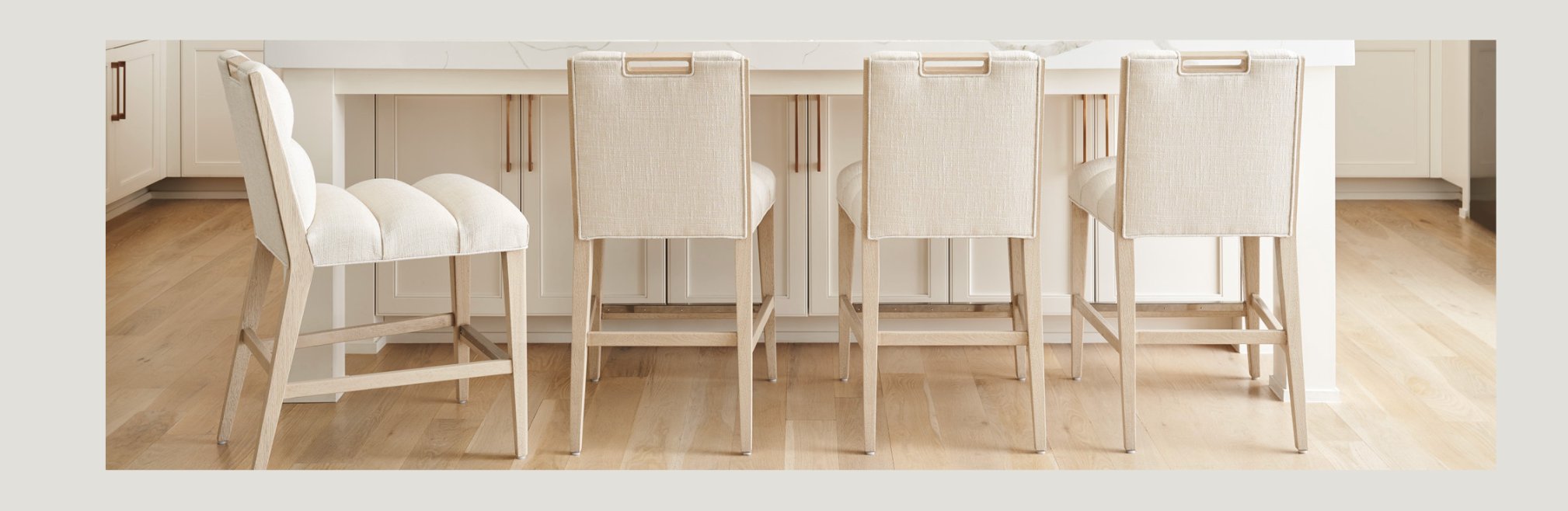 Row of upholstered counter stools with light wood frames and brass handle accents, arranged at a white kitchen island with light wood flooring