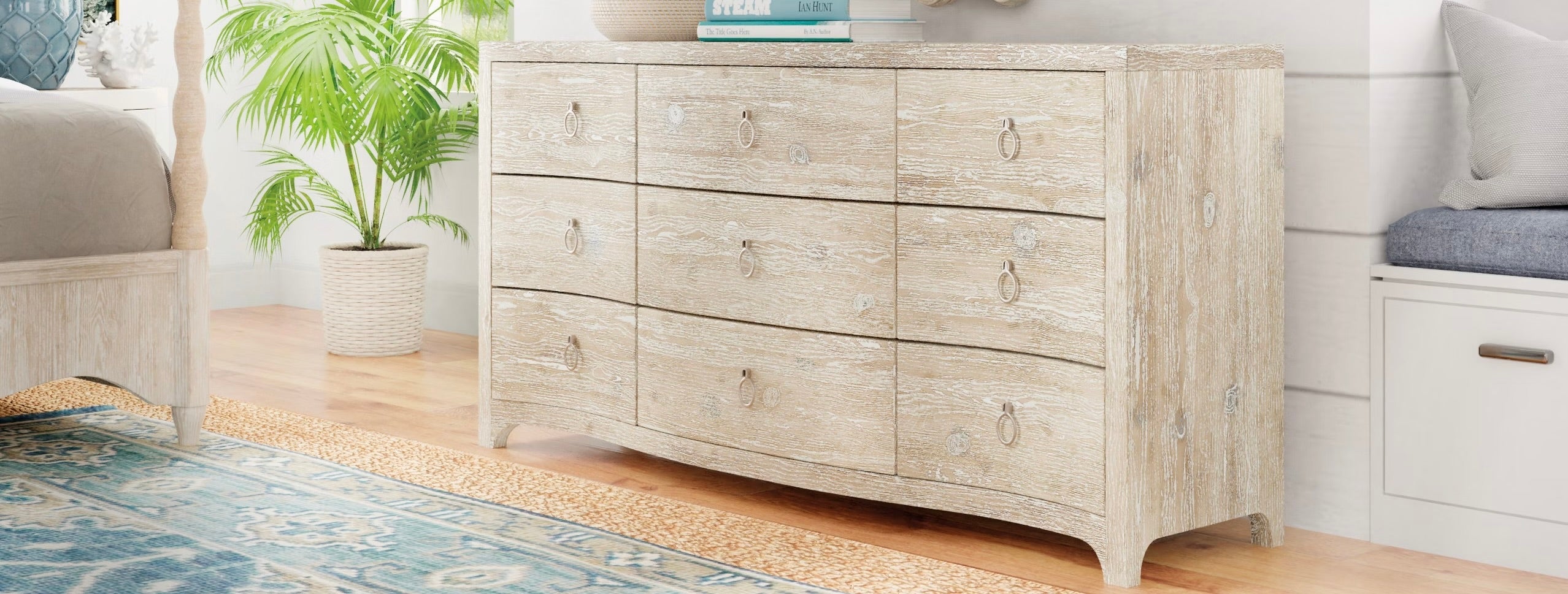 Light wood dresser with nine drawers and curved base, styled in a coastal bedroom with a blue patterned rug, potted palm, and neutral decor