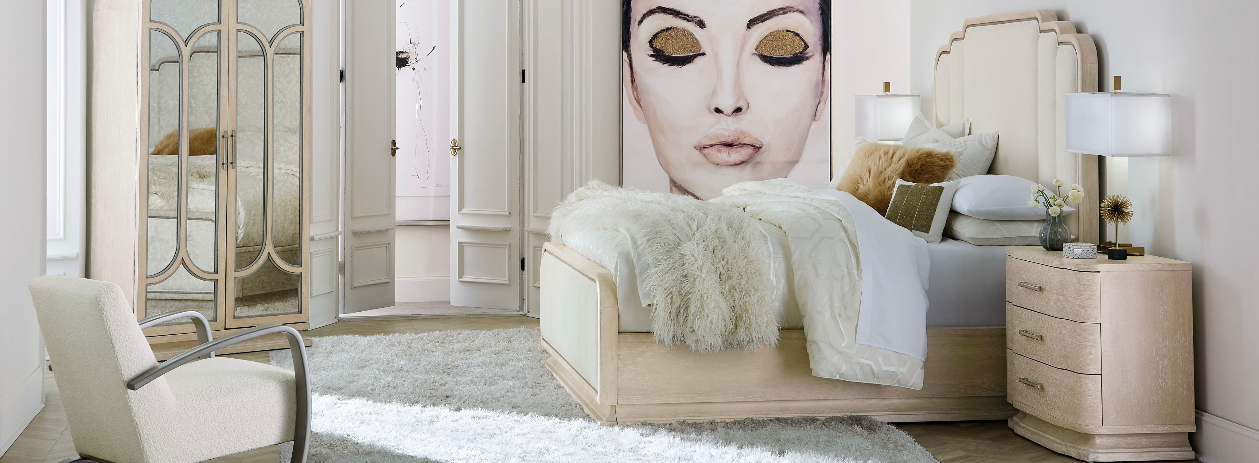 Elegant bedroom with a cream upholstered bed, matching nightstands, plush rug, and a large fashion portrait, styled with soft gold accents and mirrored wardrobe doors