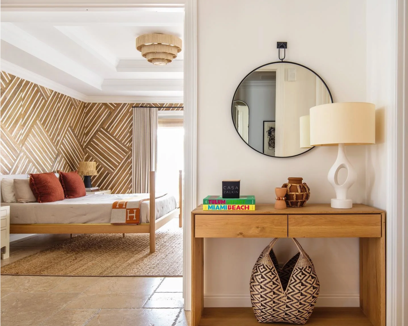 Modern bedroom and entryway with wood console, round mirror, white table lamp, and geometric accent wall.