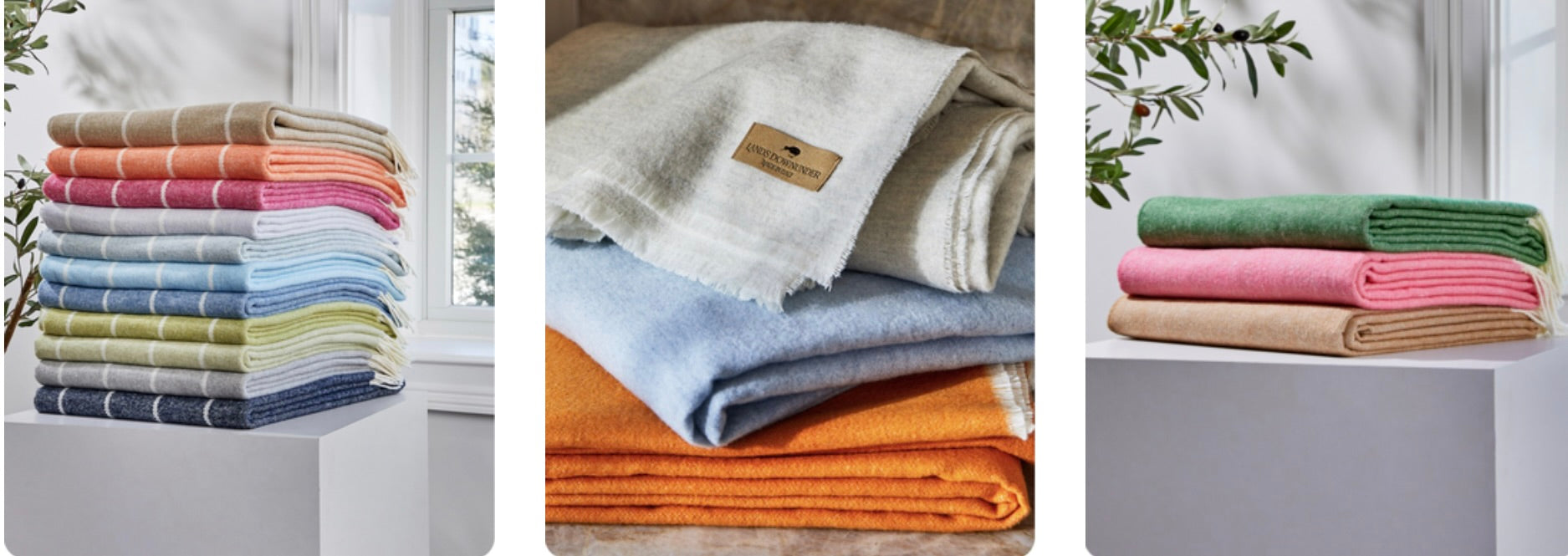 Stacked wool and cashmere throws in assorted colors including pink, green, orange, blue, and beige, styled on pedestals in a sunlit room with natural accents