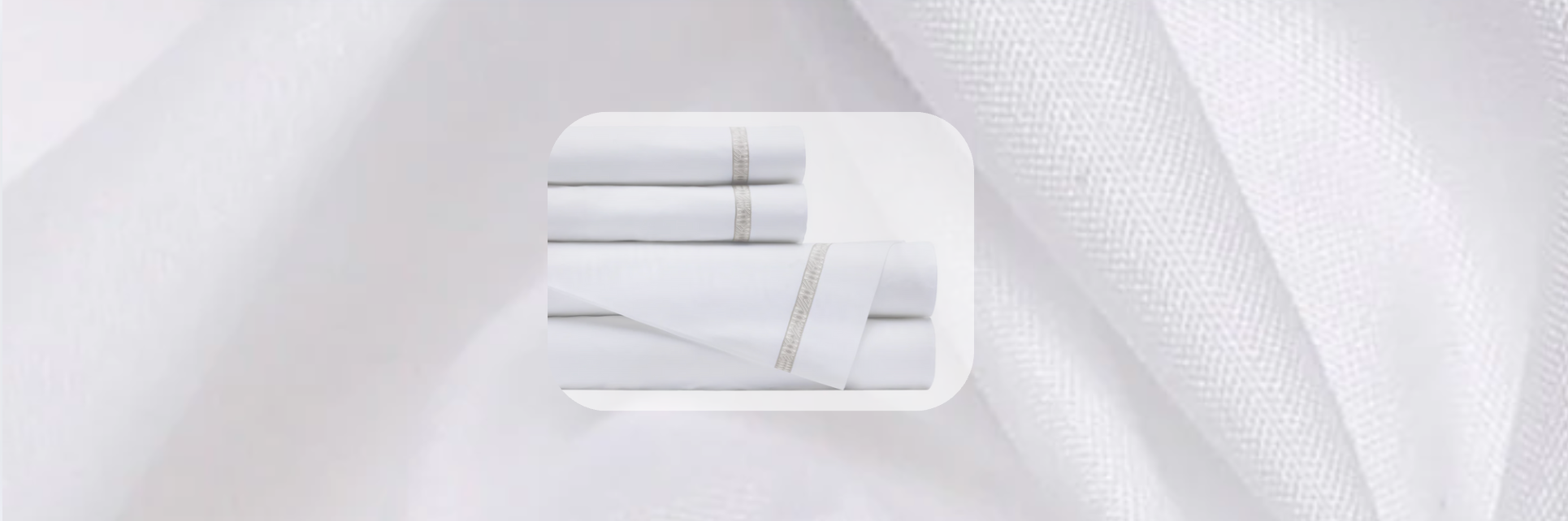 White luxury bed sheet set with embroidered border detail, folded against a soft textured fabric background